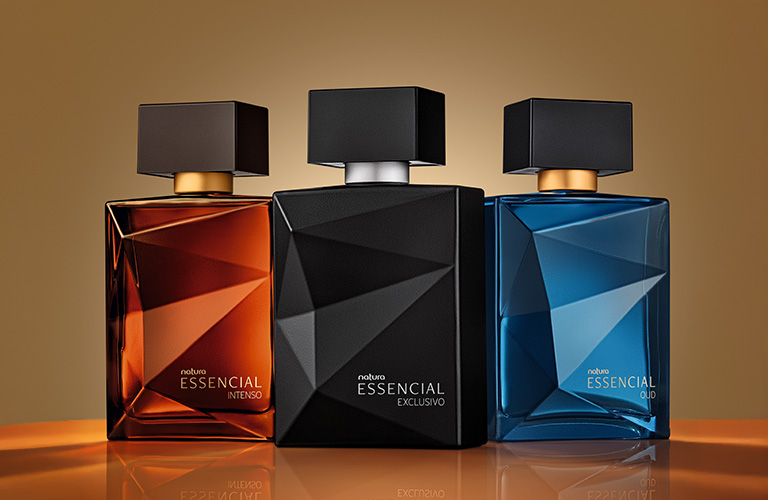 Foto de tres frascos de perfume de la línea Essencial Natura. El fondo es de color dorado uniforme, y los frascos están alineados: uno en tonos ámbar, otro negro y el tercero azul, sobre una superficie reflectante.